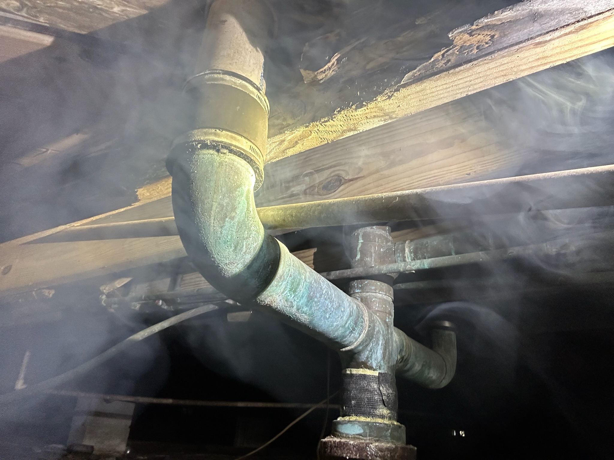 Sewer smoke escaping from connections between cast iron, copper, and PVC pipes under a house during leak detection. 📄 Description: