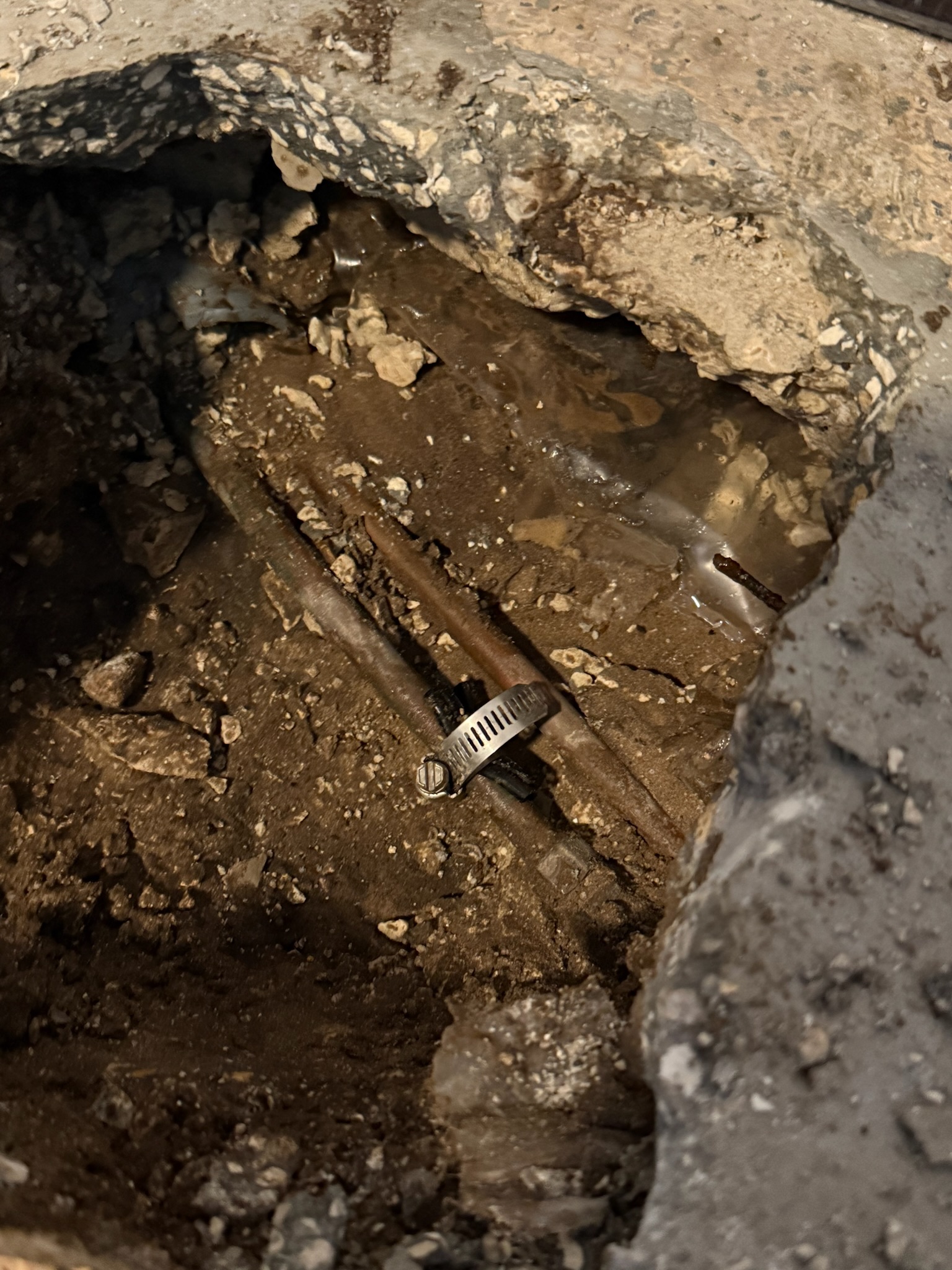 Close-up of two copper pipes under slab, one with clamp repair and surrounding sand