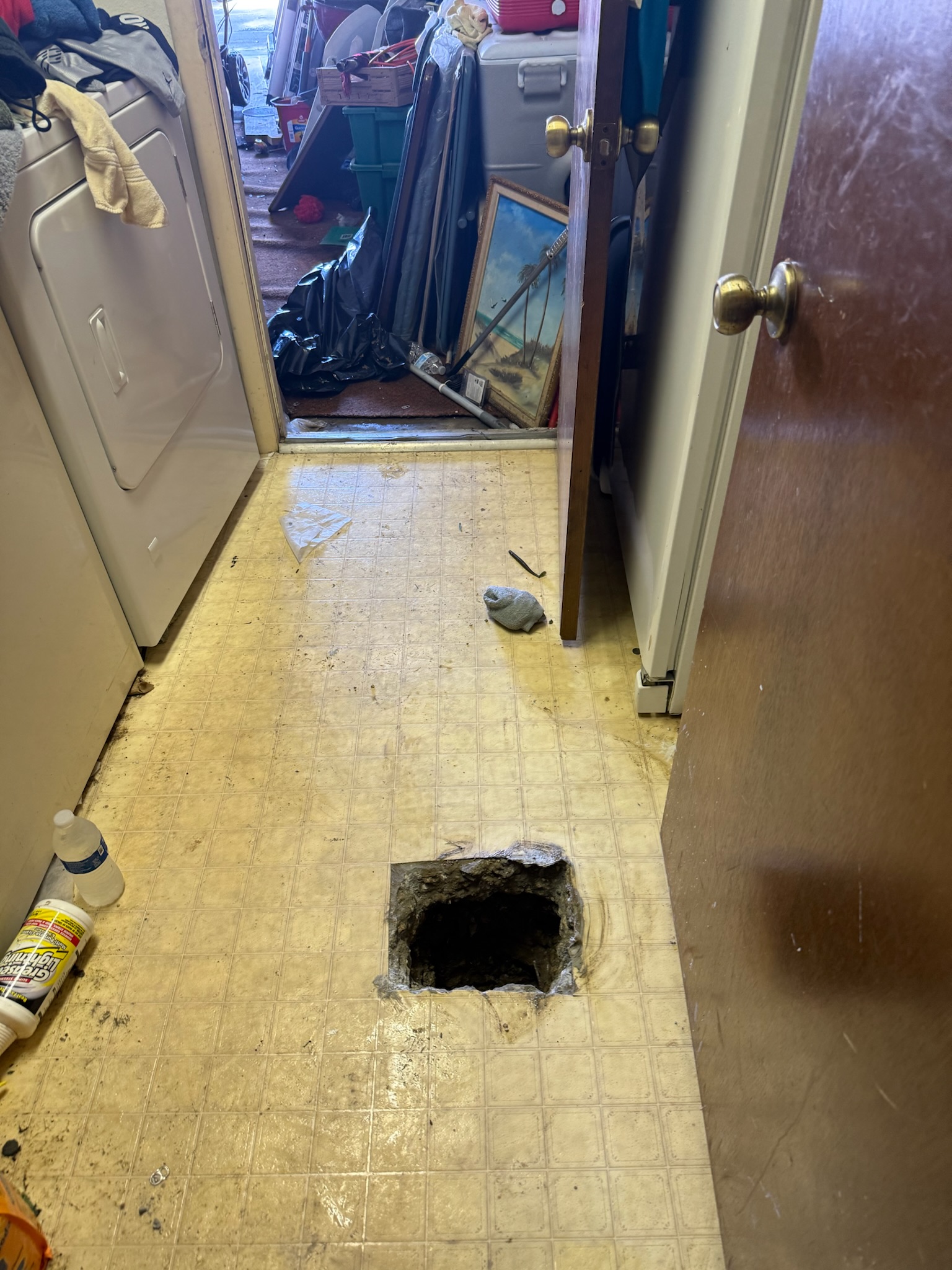Cluttered laundry area with open door, items piled on dryer, and square cut in floor exposing slab leak
