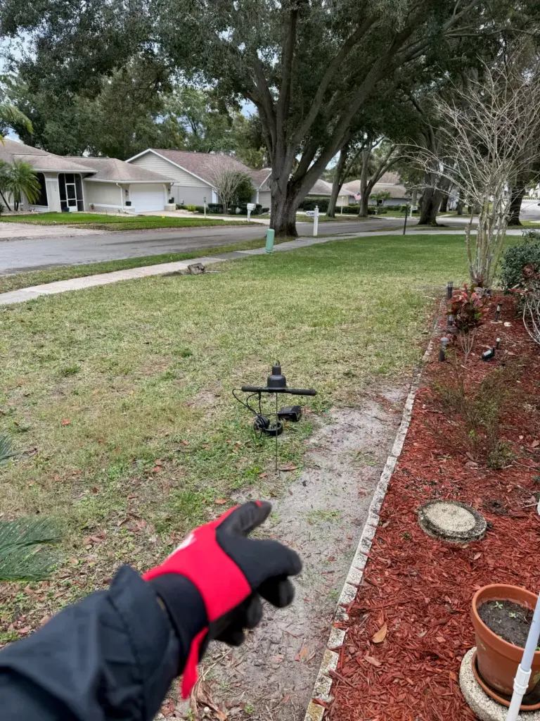 Gloved hand pointing to an acoustic listening microphone set on a lawn beside a mulched flower bed.