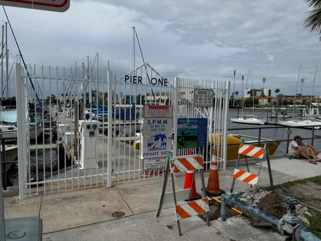 Orange barricades and a cone around an excavated water line beside a white marina gate.