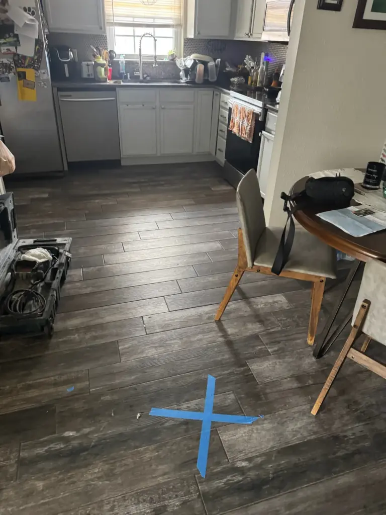 Blue painter's tape X marking a spot on a dark wood-look tile floor near a kitchen and dining table.