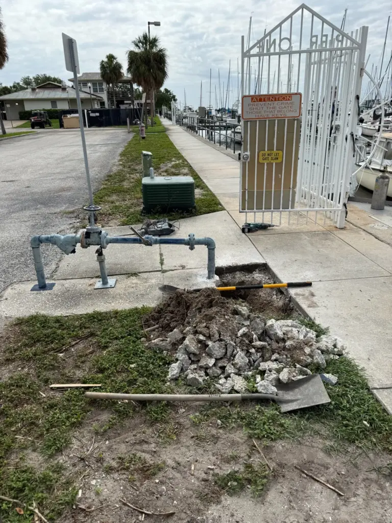 Blue pipe riser assembly beside a dug hole, a pile of broken concrete and a shovel on the grass near a marina.