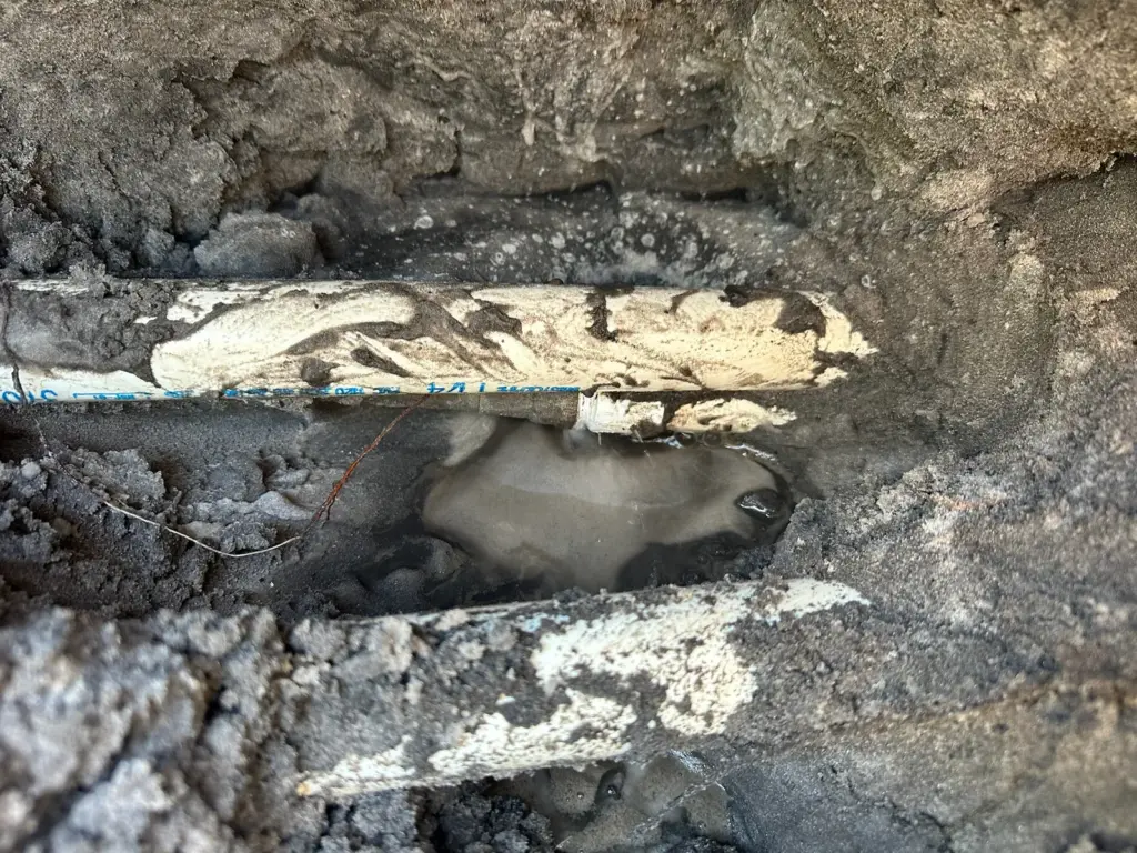 Two PVC pipes exposed in a dug hole of sandy soil, with muddy water pooled beneath them.
