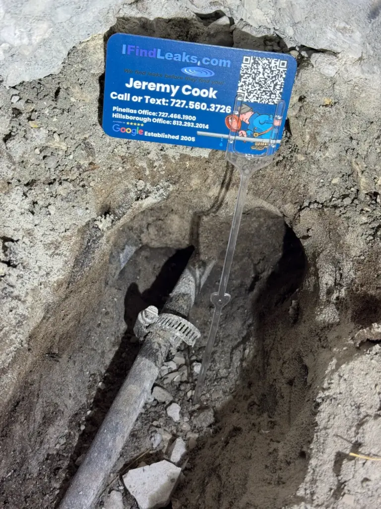 An I Find Leaks business card on a clear stake beside a gray pipe with a hose clamp in a dug hole.