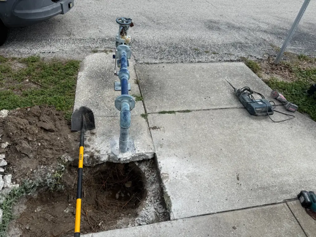 Concrete pad broken out beside a blue water pipe riser, with a dug hole, shovel and rotary hammer nearby.