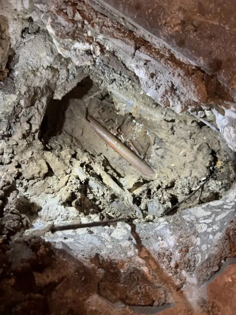 Copper pipe exposed in wet soil inside a rough opening broken through a concrete slab.