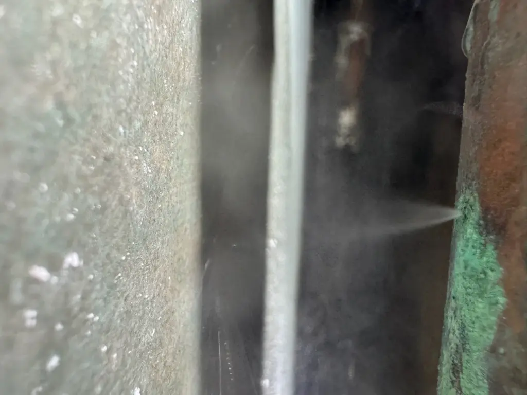 A fine mist of water sprays from a pinhole in a copper pipe crusted with green corrosion inside a wall.
