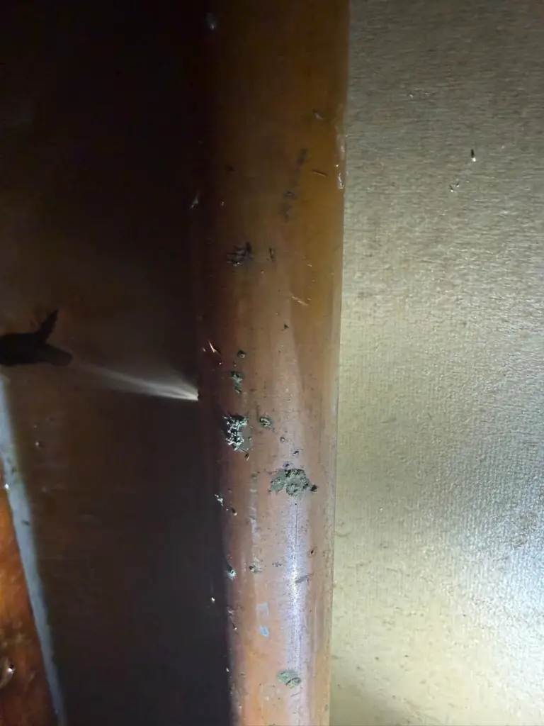 Fine spray of water shooting sideways from a pinhole in a corroded copper pipe lit by a flashlight.