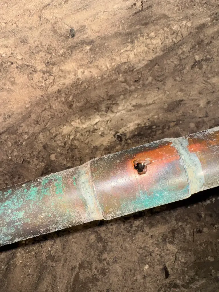 Copper pipe with green corrosion and a small dark pinhole beside a soldered joint, lying in soil under a slab.