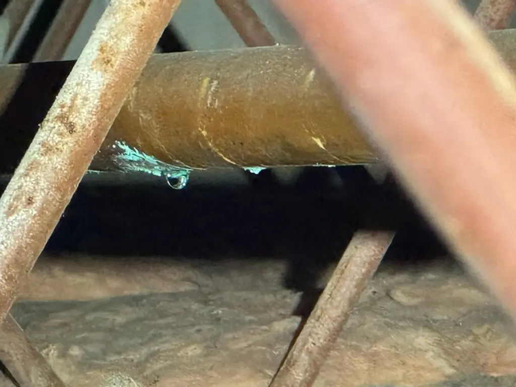 A water drop hangs from a spot of green corrosion on a copper pipe running between rusty trusses.