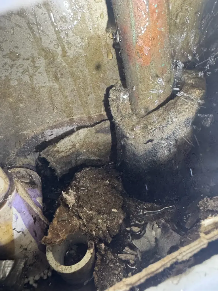 Wet, corroded copper pipe coming up through broken concrete next to a white drain stub and loose debris.