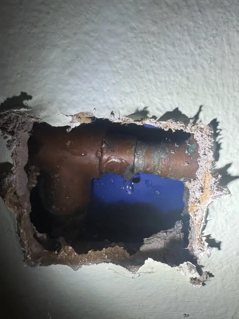 A rough hole cut in textured drywall, exposing copper pipes with a strip of blue tape inside.