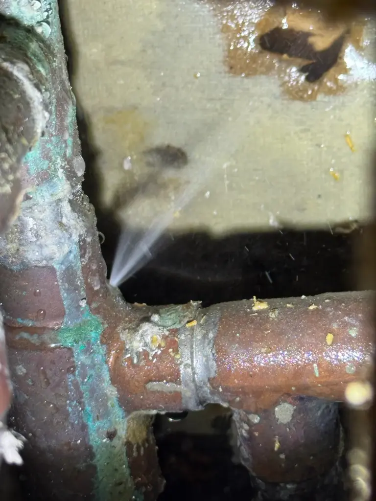 Thin jet of water spraying from a pinhole in a corroded copper tee with green patina.