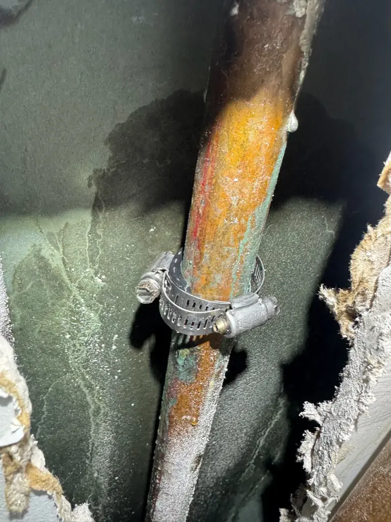 A corroded copper pipe with orange and green staining and a metal hose clamp inside an opened wall cavity.