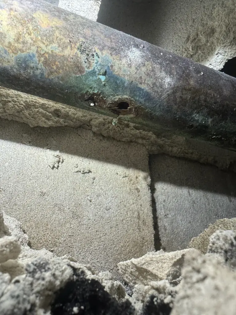 Discolored, corroded copper pipe with a small hole eaten through it, resting on concrete block in an opening.