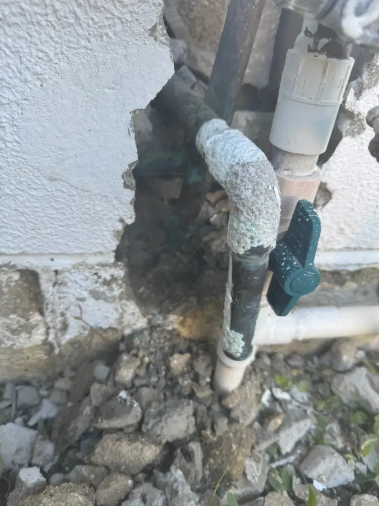 Corroded copper pipes and a green shutoff valve exposed in a hole broken through an exterior block wall.