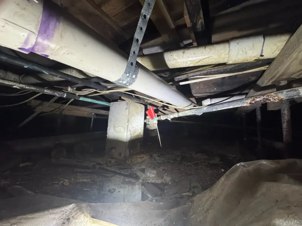 Wide view under a raised floor with white drain pipes overhead and a red flag marking a supply pipe.
