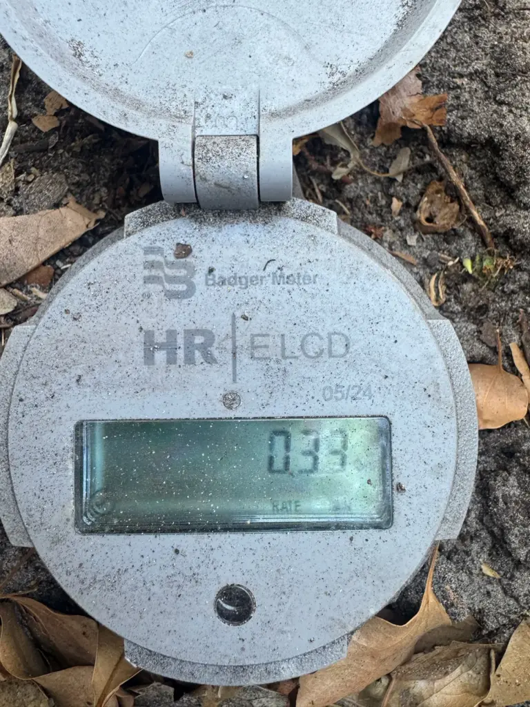 A digital water meter with its lid flipped open shows a reading on its display among dry leaves.