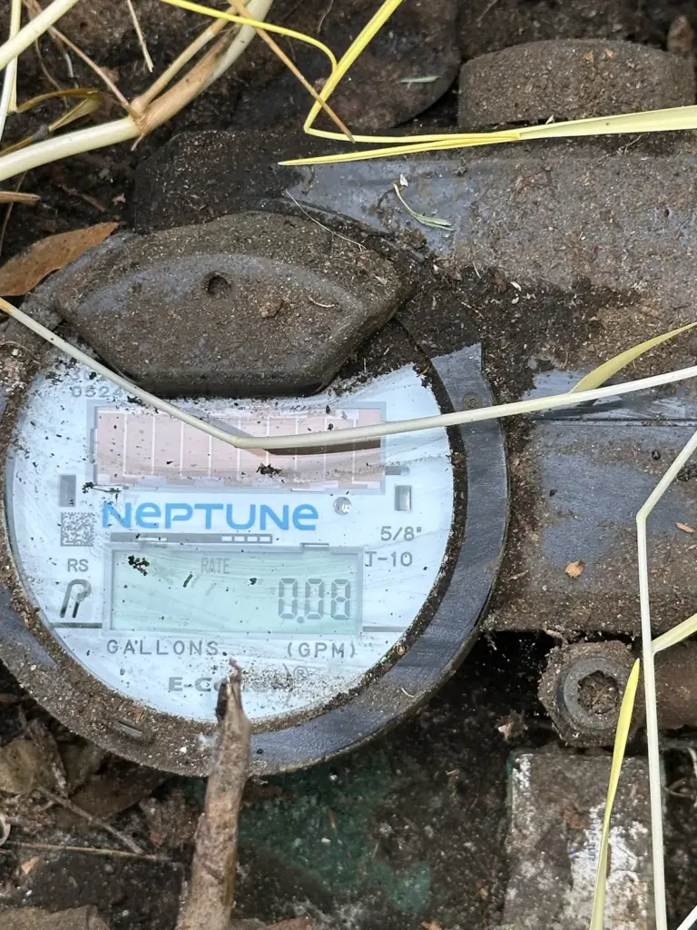 A digital water meter inside a meter box shows a flow reading on its display, surrounded by dirt and grass.
