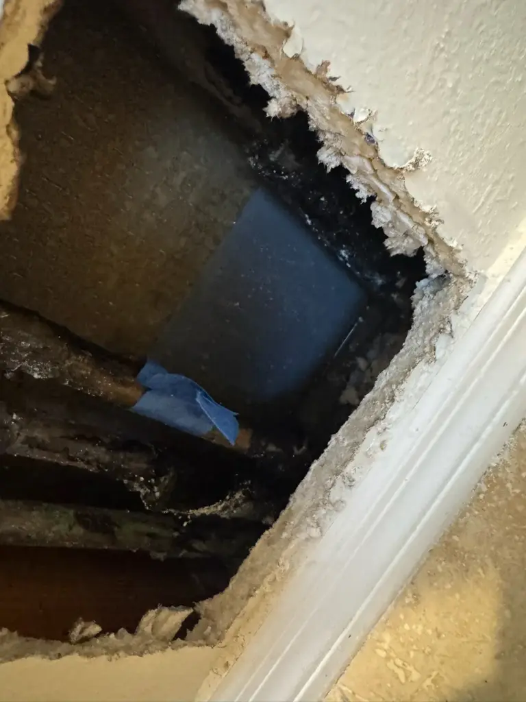 Hole cut in drywall above a baseboard, exposing a dark cavity with a pipe and blue tape inside.