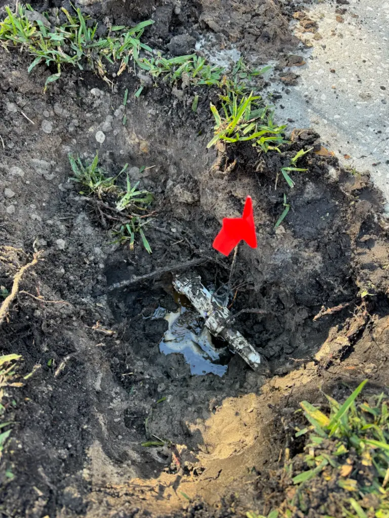 Red marker flag beside a muddy hole in a yard where a white pipe is exposed in standing water.