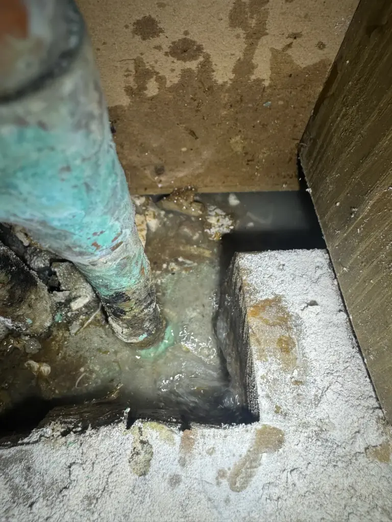 Close view of a copper pipe with heavy green corrosion standing in muddy water inside a slab opening.