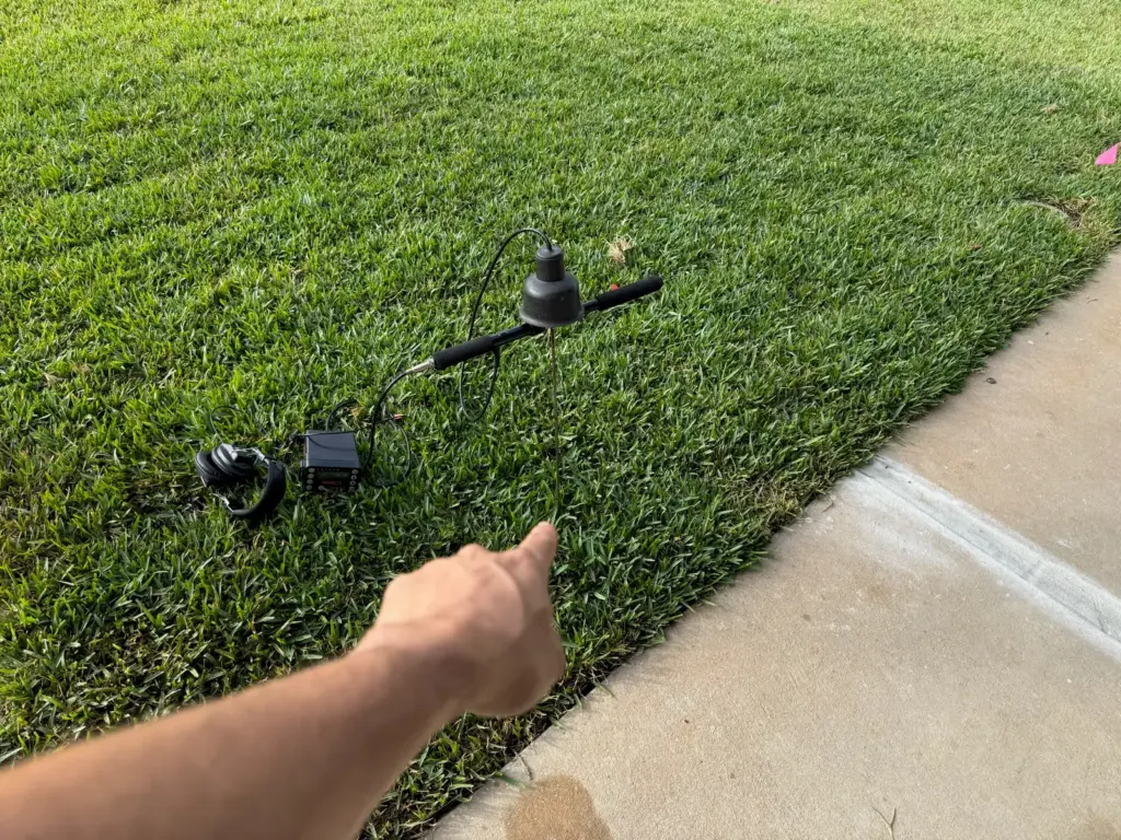 A hand places an acoustic ground microphone on a green lawn beside a concrete walkway.