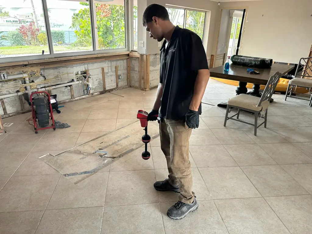 Technician holds a red pipe locator over a tile floor in a gutted room, a sewer camera reel by the windows.