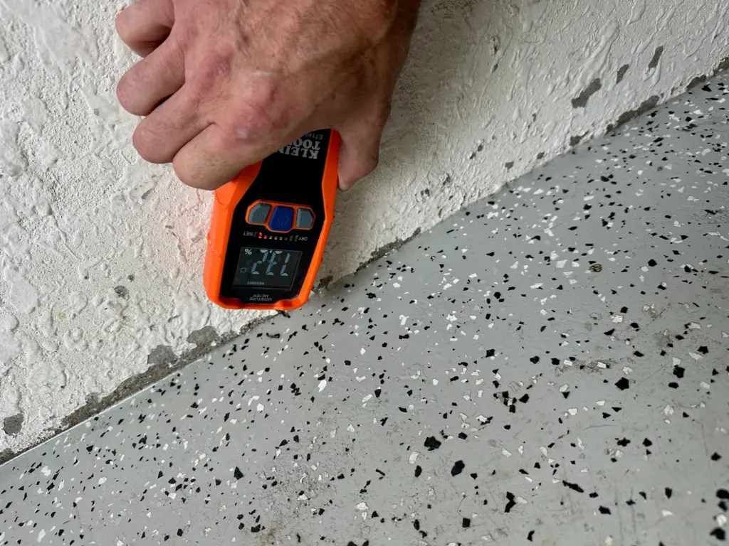 Close-up of an orange moisture meter with a lit display held at the base of a white wall above a speckled floor.