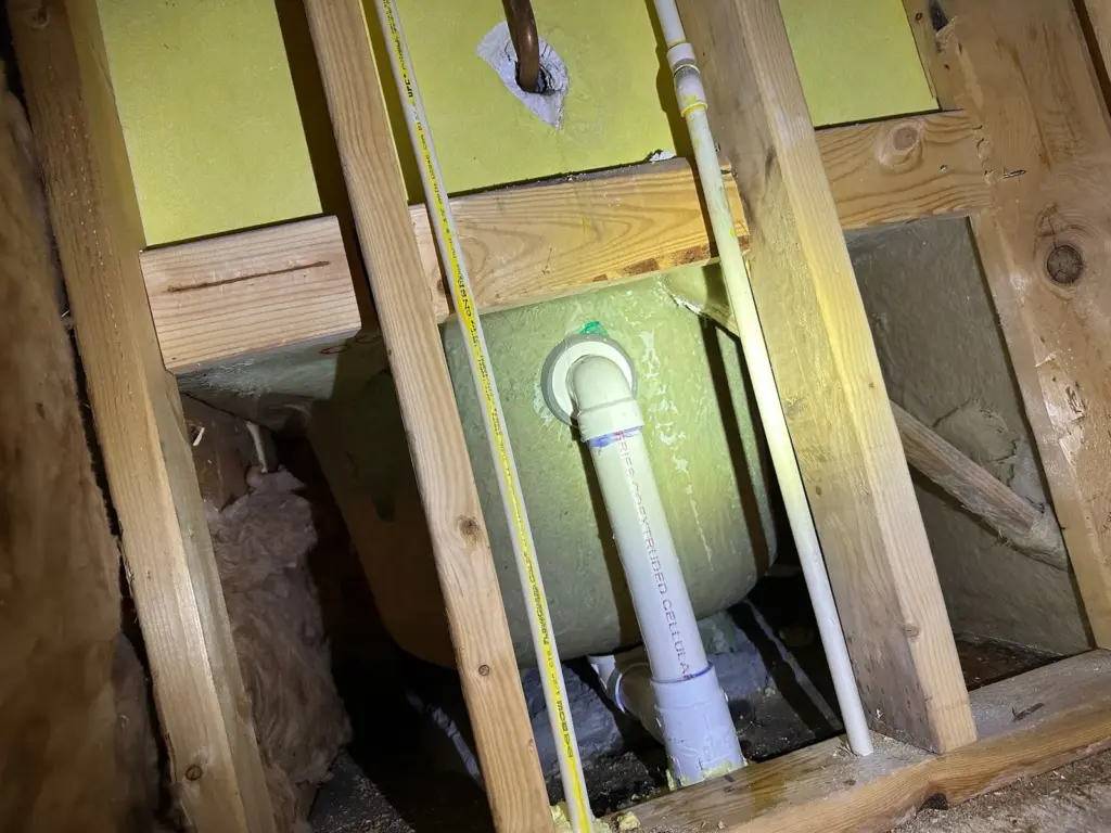 Opened wall behind a bathtub showing a white drain pipe, CPVC water lines and wood framing.