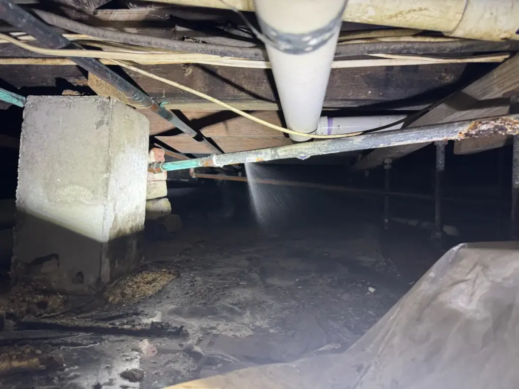 Fine spray of water shooting from a corroded supply pipe beneath floor joists in a dark crawlspace.
