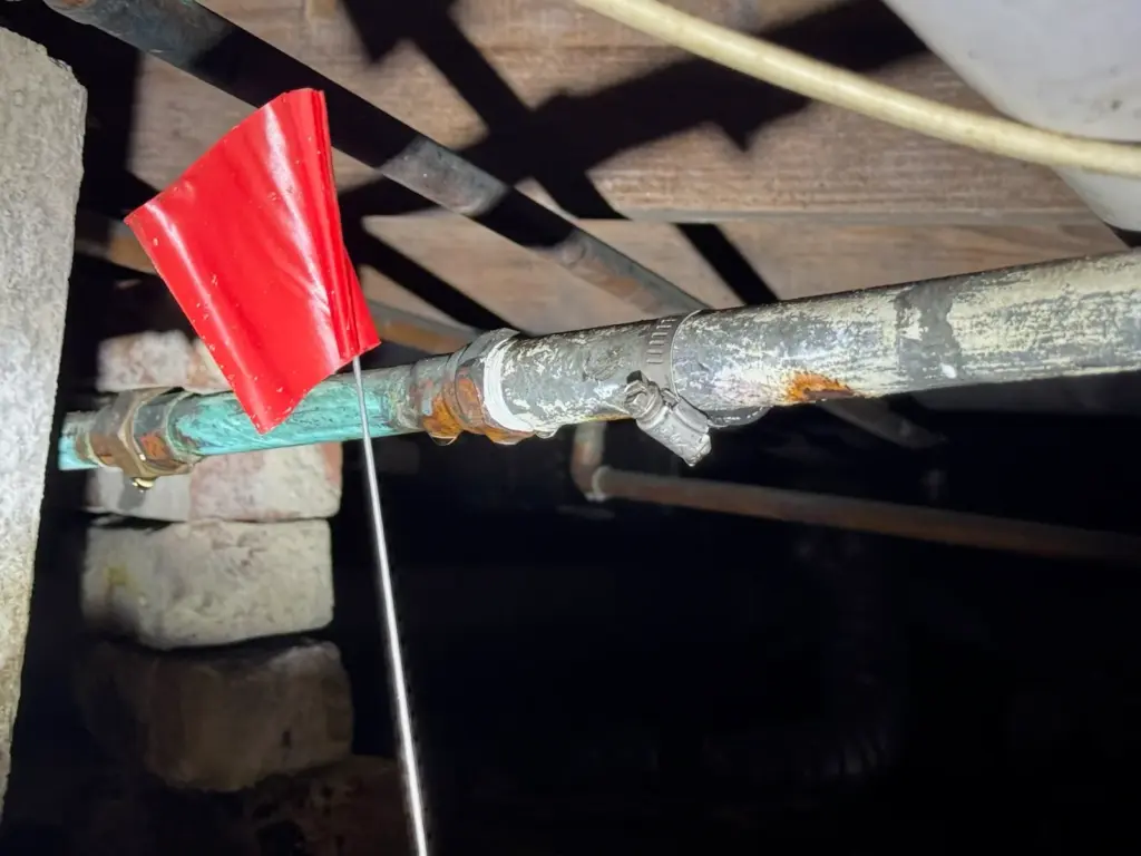 Red marker flag set beside a corroded pipe fitting under the floor joists of a crawlspace.