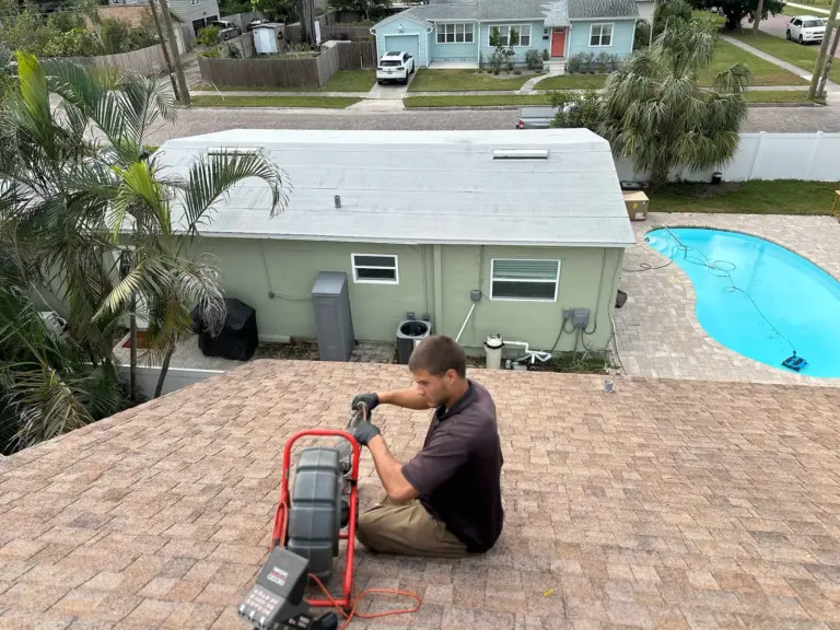 Technician seated on a shingle roof feeding a sewer camera from a red reel, with a pool and neighborhood behind him.