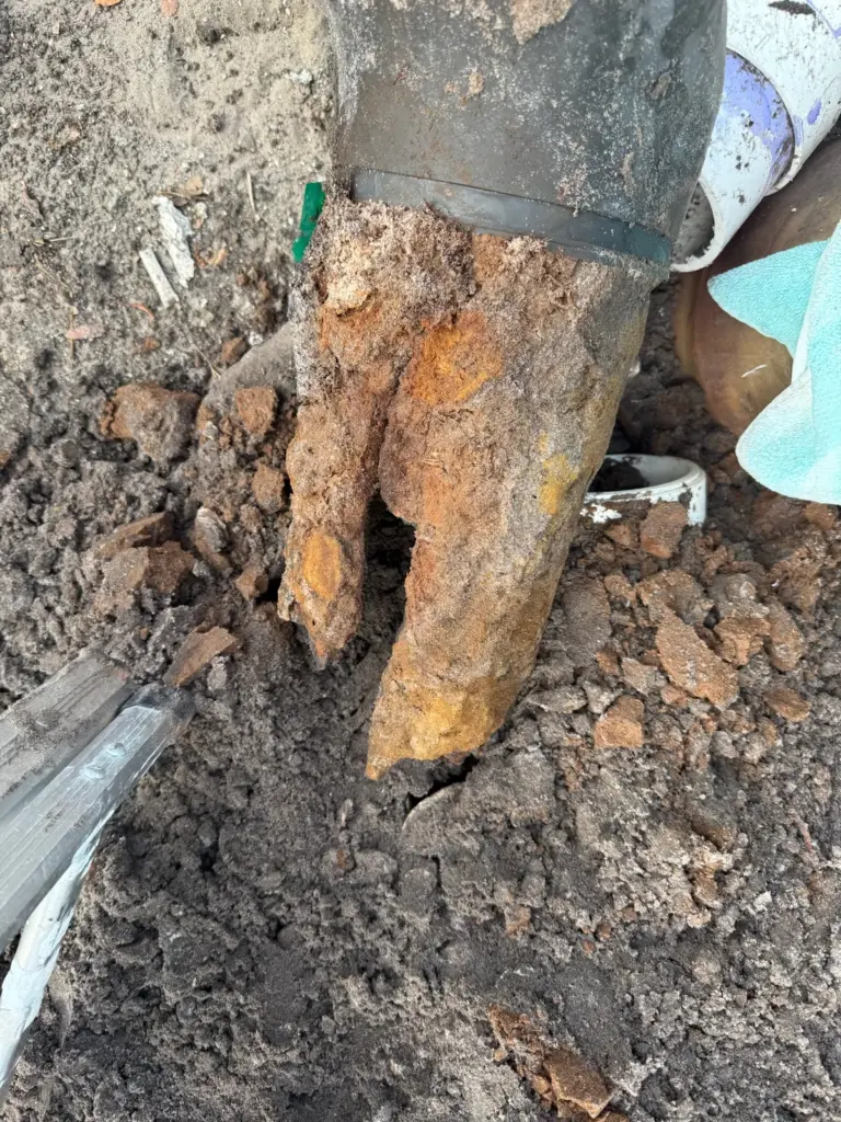 Rusted cast iron pipe end split apart and crumbling, exposed in dug soil beside a rubber coupling.