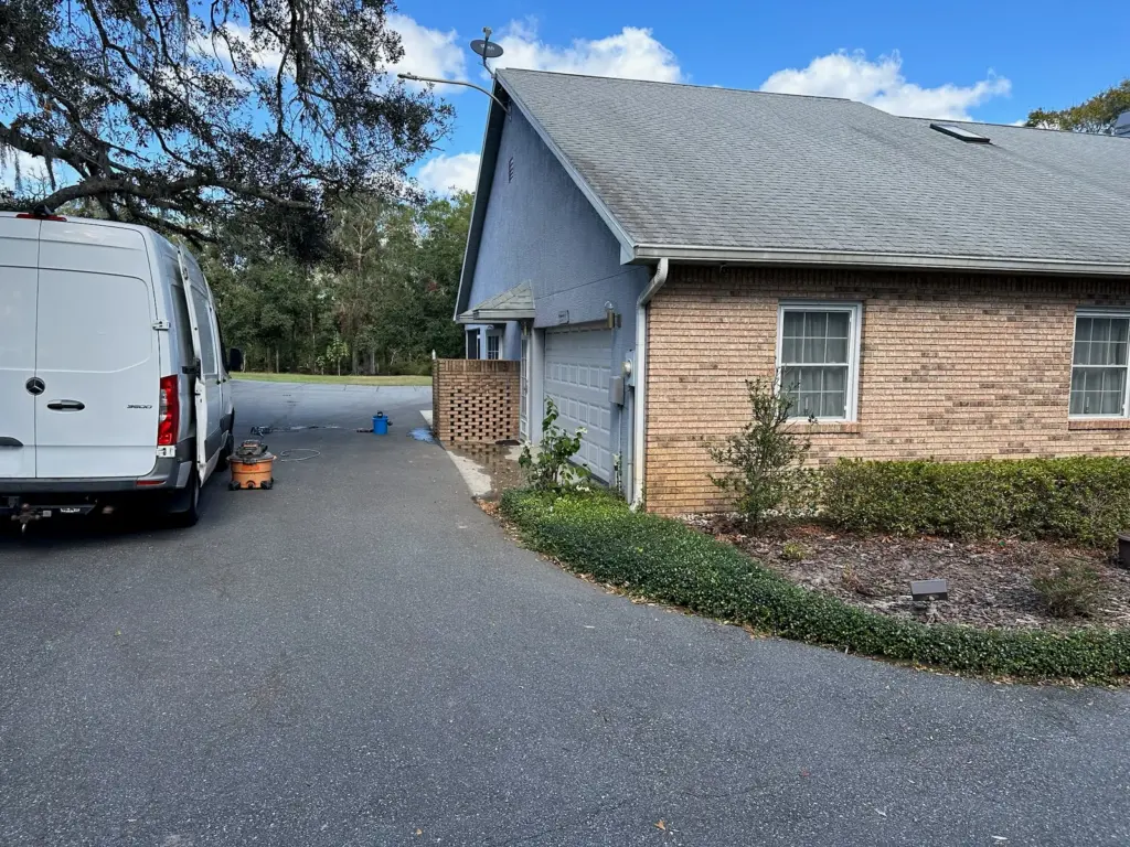 White service van parked in a driveway beside a brick house, with a shop vacuum and a bucket set out on the pavement.