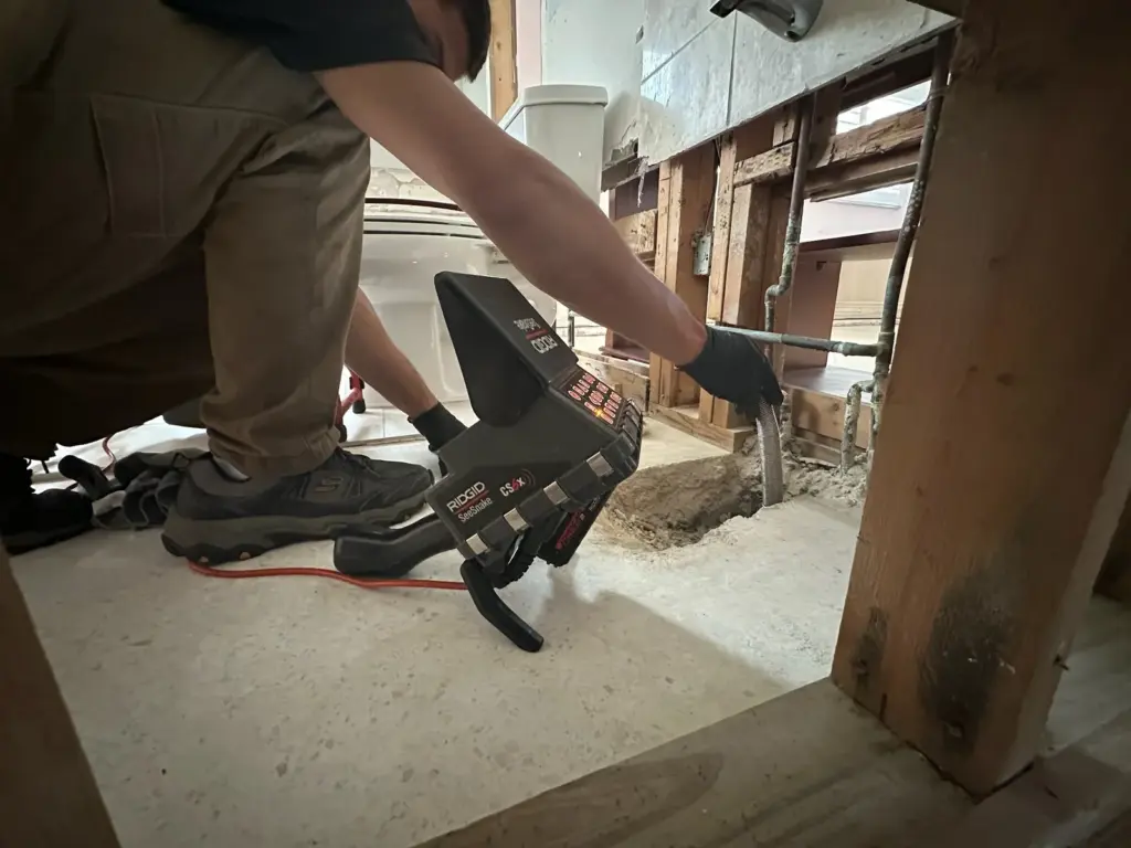 Technician feeds a camera cable into an opening dug in the floor beside a sewer camera monitor.