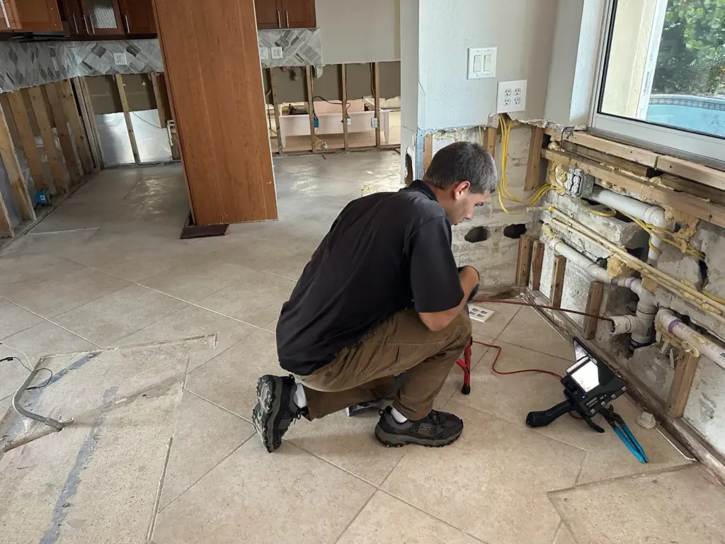 Technician kneels at exposed plumbing in a gutted room with drywall removed, a camera monitor beside him.