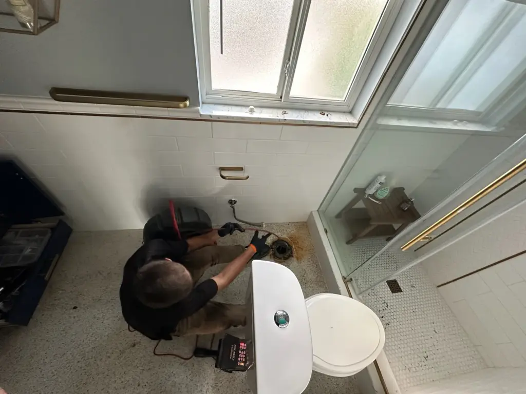 Overhead view of a technician feeding a sewer camera cable into an open toilet drain on a bathroom floor.