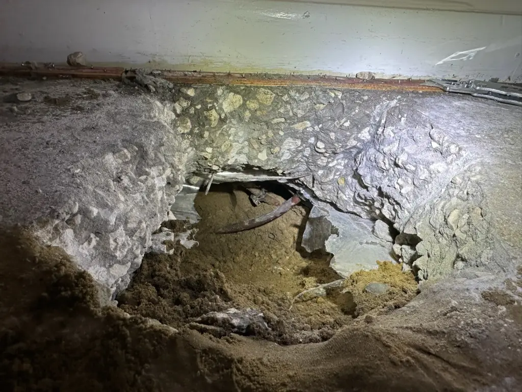 Large hole broken in a concrete slab along a wall base, exposing sandy soil and a copper pipe.