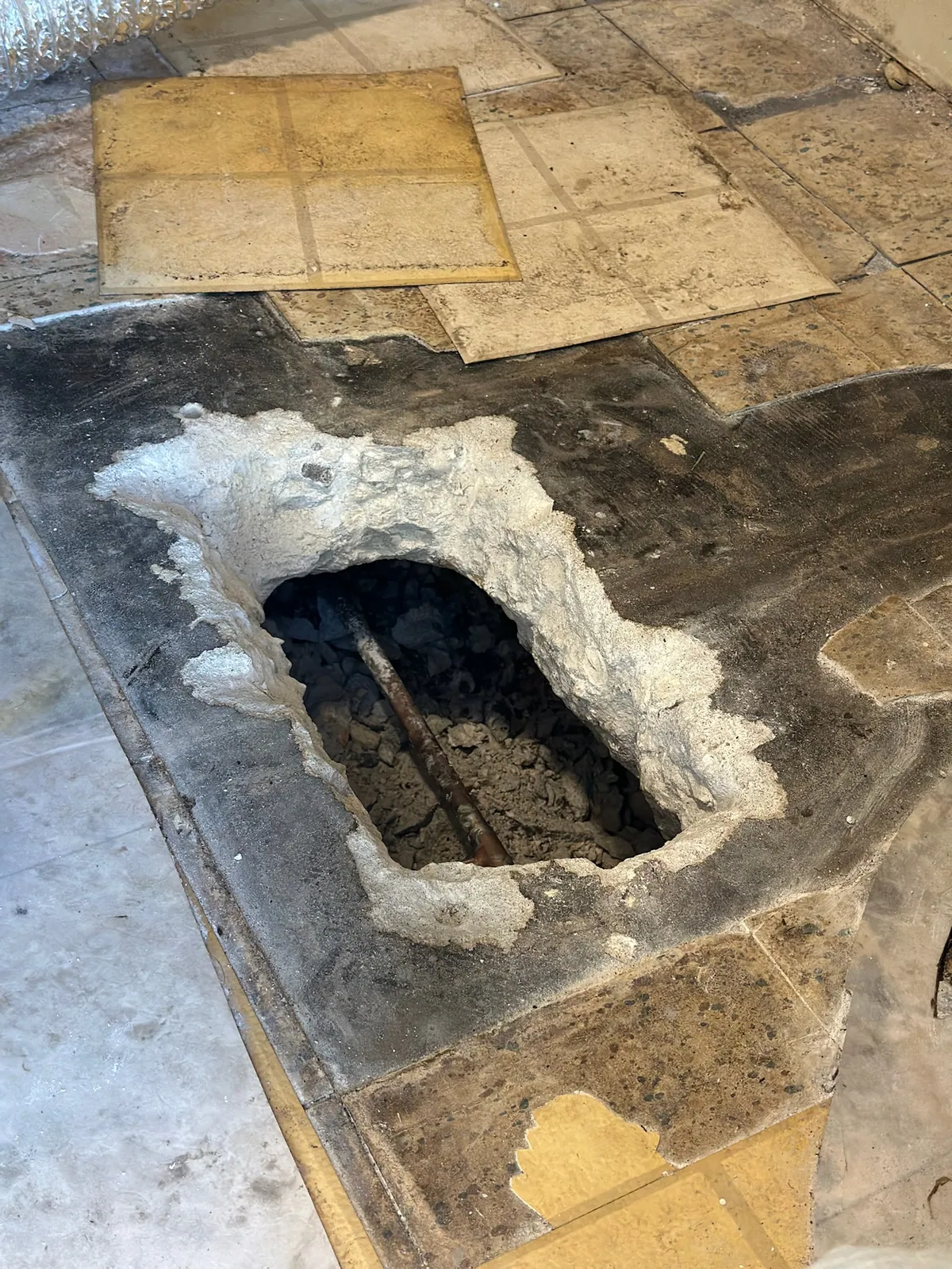 Opening broken through a concrete floor where tiles were removed, exposing pipes in the soil below.