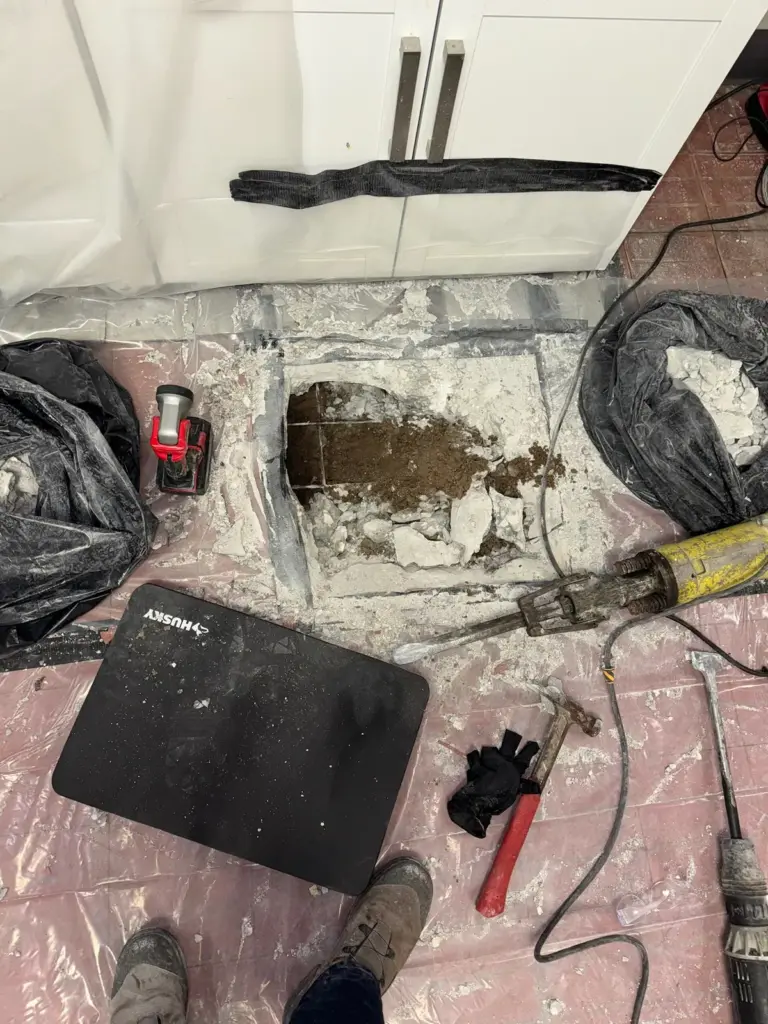 Opening broken through a kitchen slab with a jackhammer chisel, hammer, knee pad and gloves on plastic sheeting.