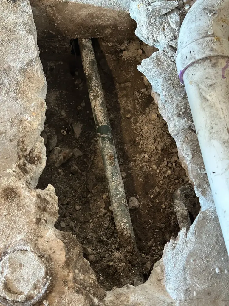 Long trench broken through a concrete slab, exposing a pipe running through soil beside a white drain pipe.