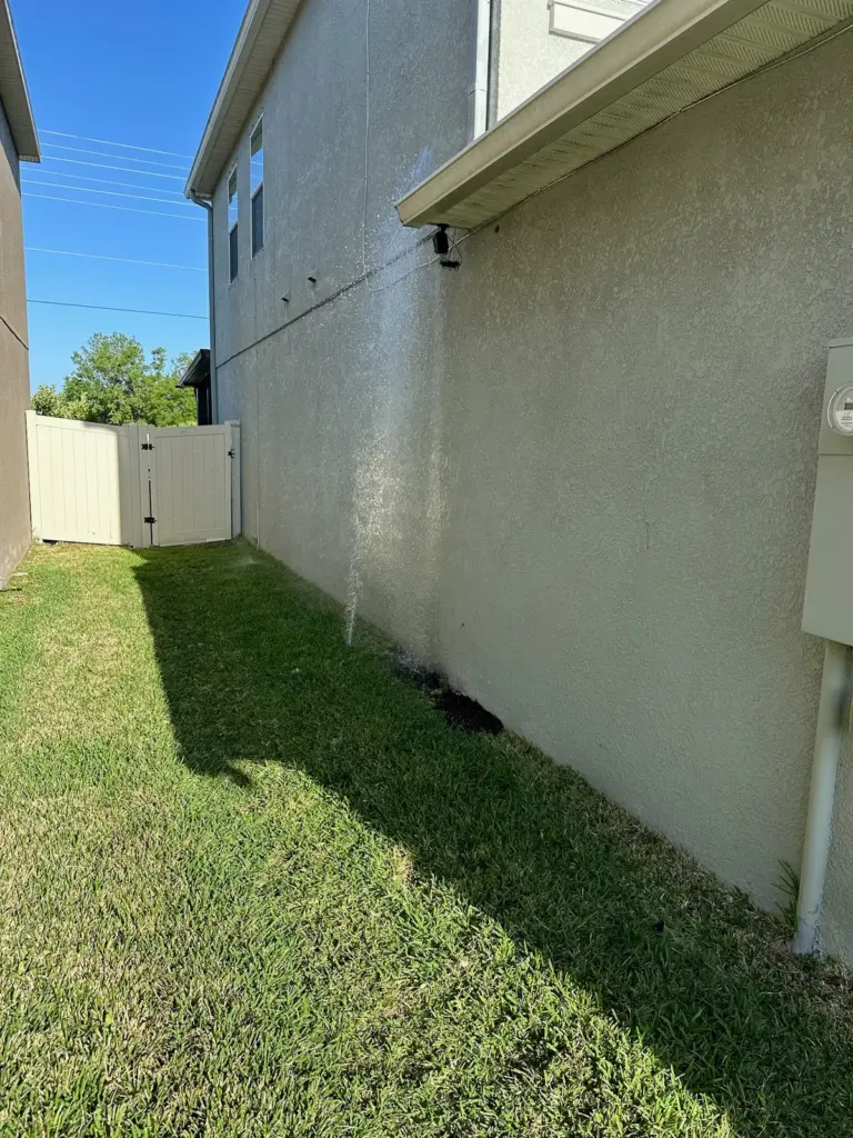 A lawn sprinkler sprays water against the stucco wall of a two-story house in a narrow side yard.