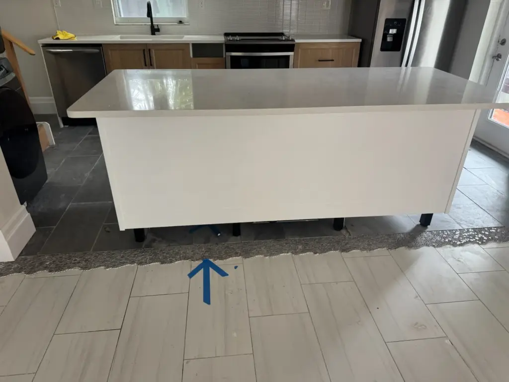 White kitchen island with a blue tape arrow on the tile floor and a strip of flooring removed around it.