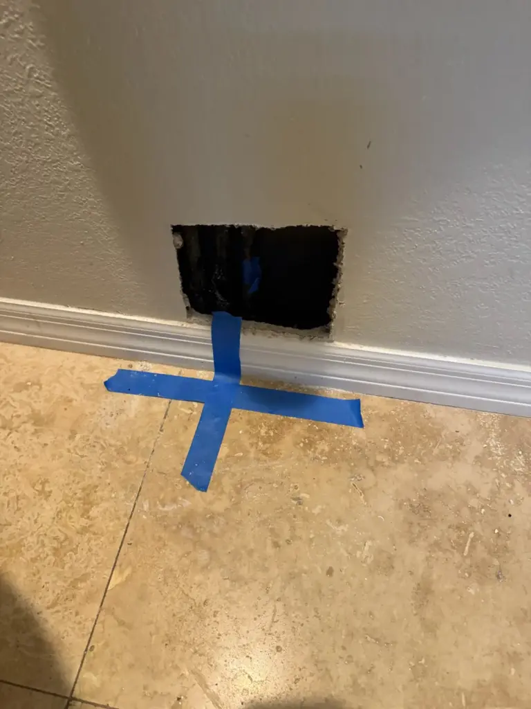 Blue tape cross on a beige tile floor directly below a square hole cut in the wall.