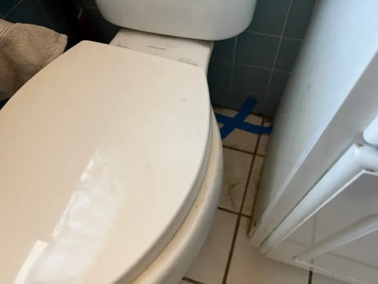 Close-up of a white toilet with strips of blue tape marked on the tile floor behind it.