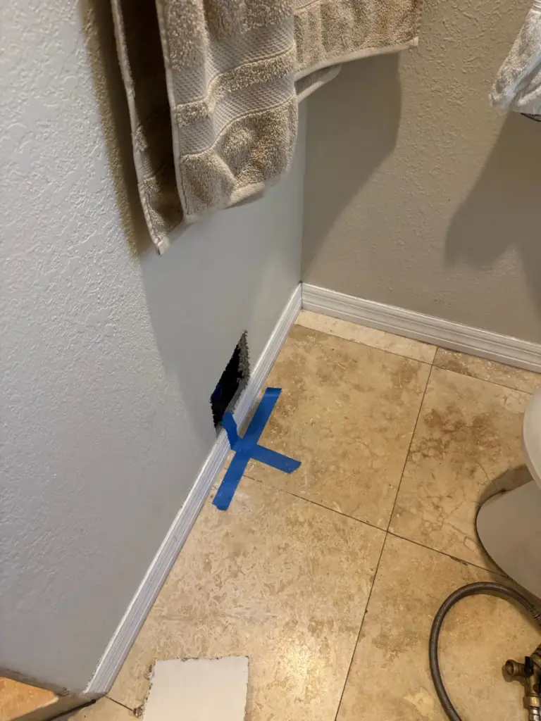 Blue tape cross on a tile floor at a bathroom wall base beside a small hole cut in the wall.