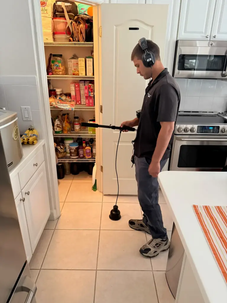 A technician wearing headphones listens with an acoustic ground microphone on a kitchen tile floor.