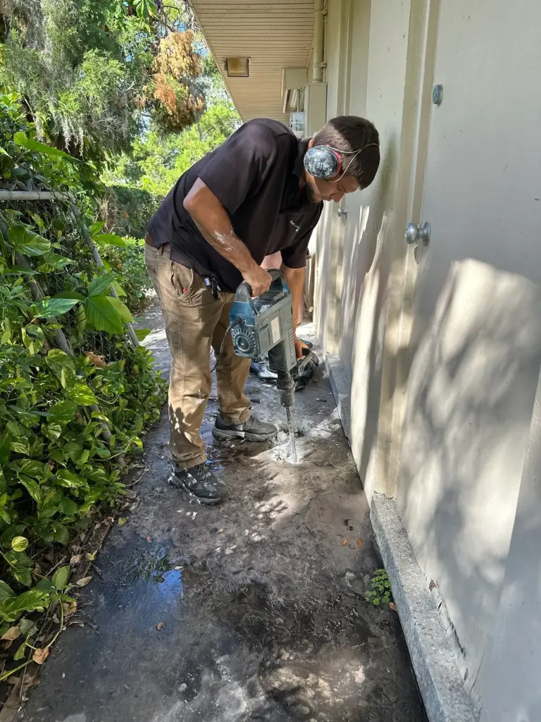 Technician wearing ear protection using a jackhammer on a narrow concrete walkway beside a house.